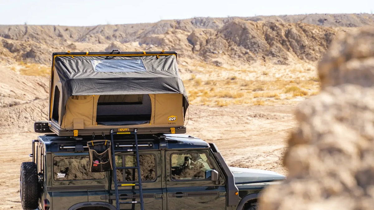 A vehicle with a rooftop tent and ladder is parked in a rocky desert landscape under a clear sky. Bushes and cliffs are visible in the background.