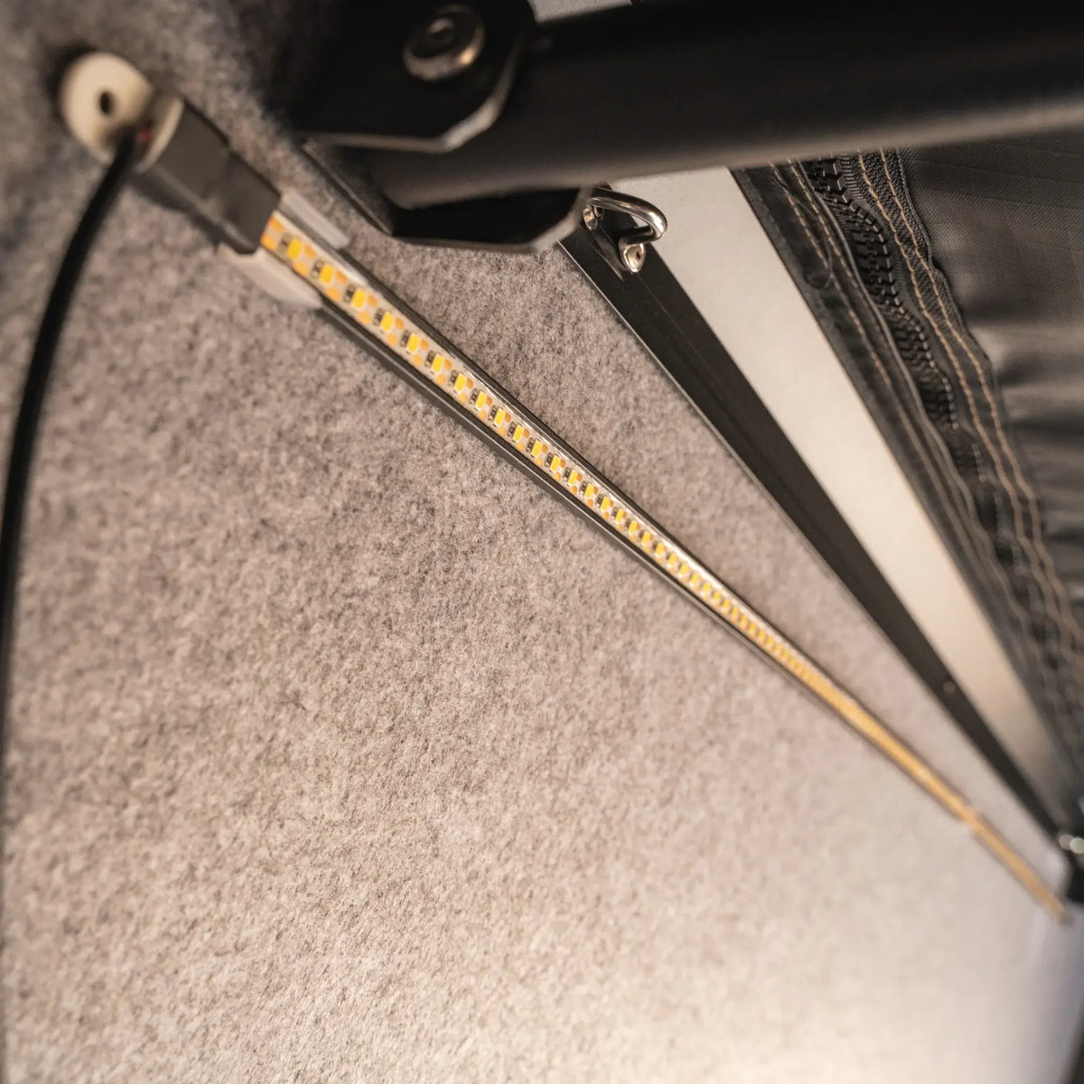 Close-up of a CVT La Sal Hardshell Tent LED strip light mounted on a gray felt surface beneath a black metal frame, with visible wiring and a zipper at the top right—highlighting the side-open design characteristic of spacious, comfort-focused rooftop tents.