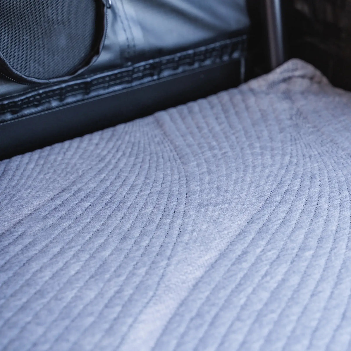 A close-up of the textured, light gray quilted fabric interior of the CVT La Sal Hardshell Tent, with a black speaker and metal frame in the background, showcases the spacious comfort and modern side-open design you can expect from this premium rooftop tent.