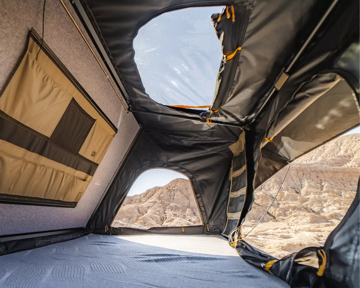 Interior view of a rooftop tent with open windows and skylight, showing a rocky desert landscape outside. The tent has fabric walls, mesh pockets, and a textured sleeping surface.