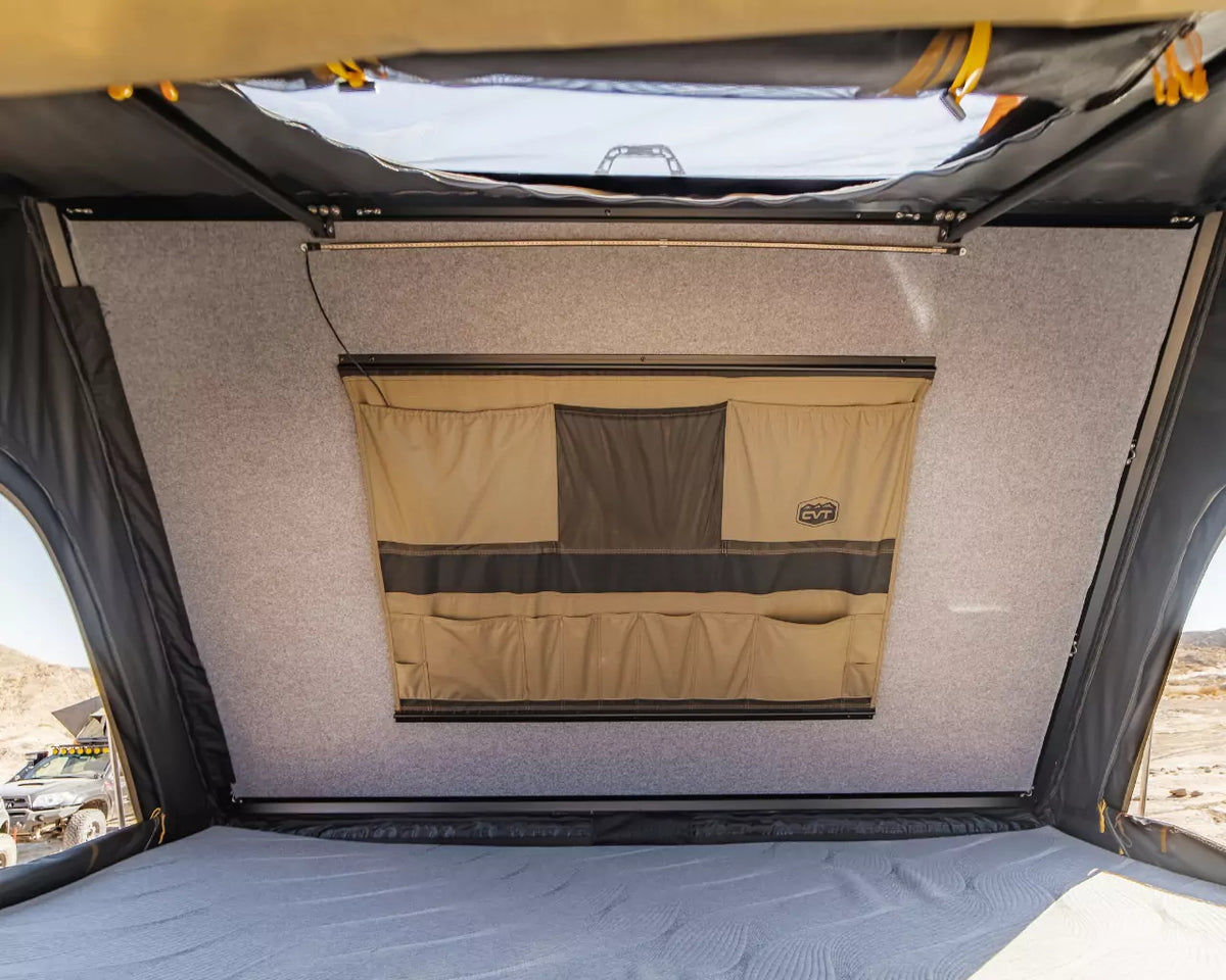 Interior view of a rooftop tent showing a closed, tan and black fabric window, a grey wall, and part of a mattress, with a desert landscape visible outside.