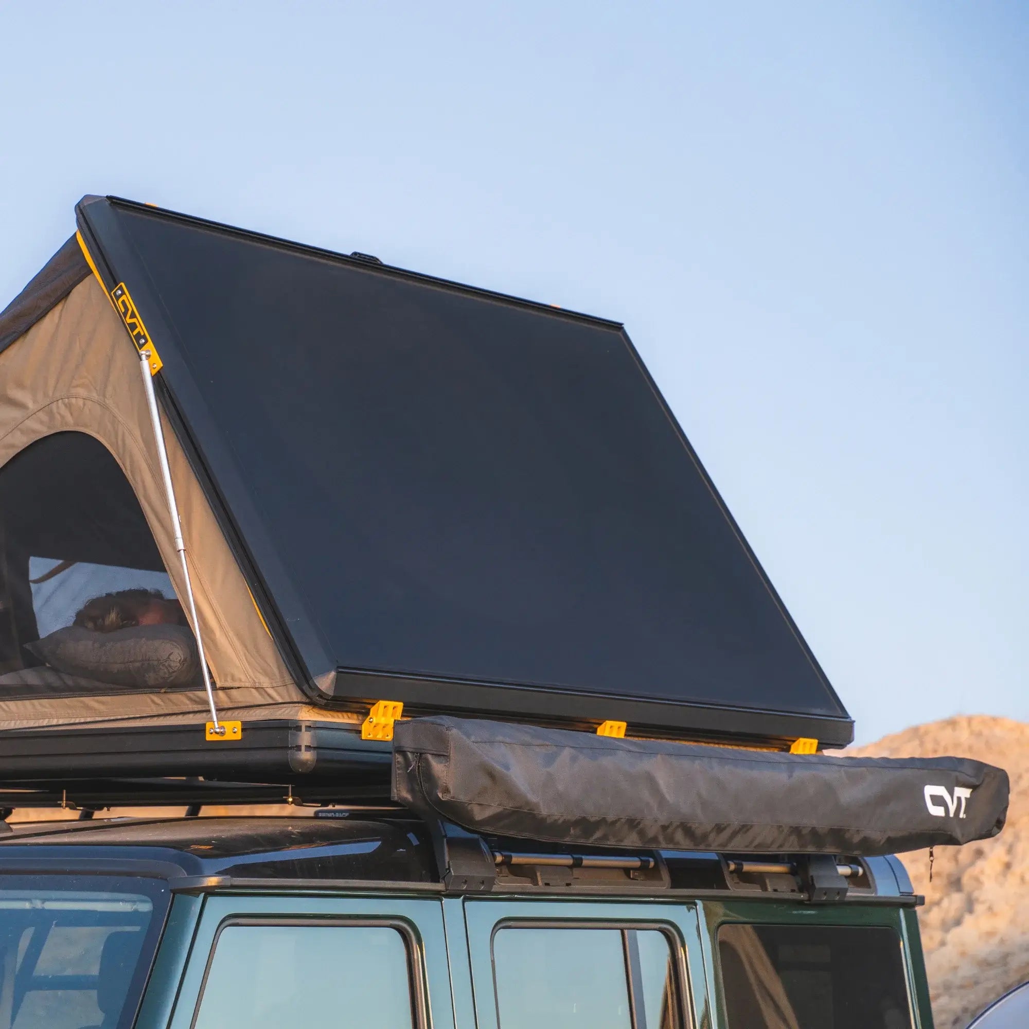 A rooftop tent with a hard shell cover is mounted on top of a vehicle. The tent is partially open, and a rolled-up awning is attached below. The background shows clear sky and rocky terrain.