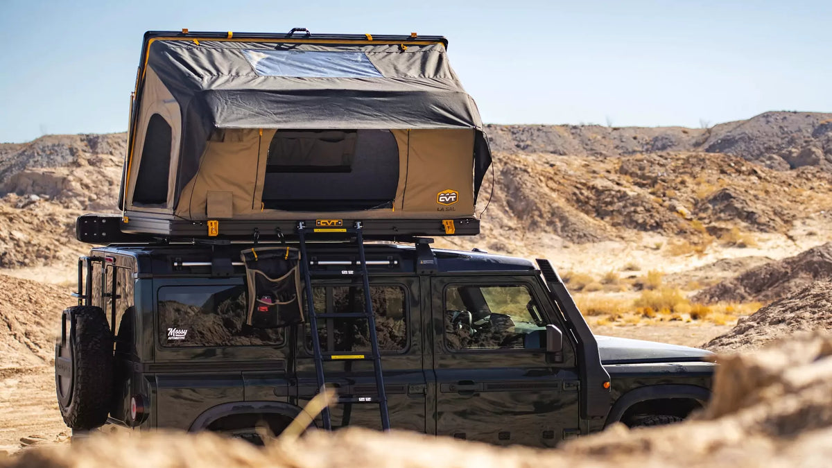 A black off-road vehicle with a rooftop tent and ladder is parked in a rocky, desert landscape under a clear blue sky.