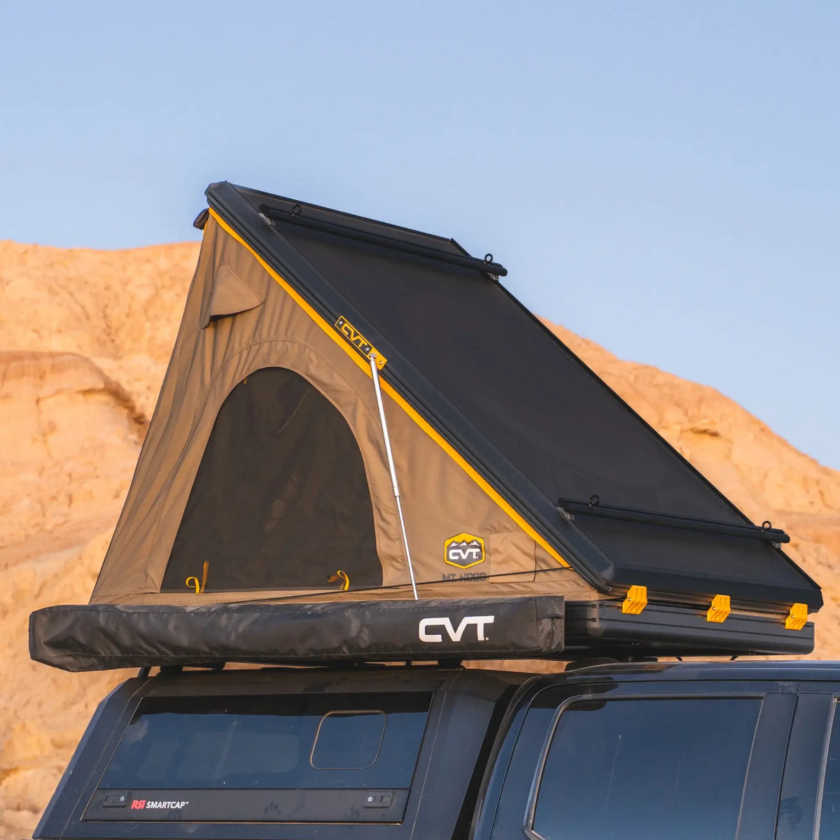 The CVT Mt. Hood/La Sal Cross Bar - 2 Pack, shown installed on top of a vehicle, serves as a durable mounting solution for a hardshell rooftop tent in tan and black with yellow accents and a mesh window. The setup is displayed with one side elevated against a rocky desert backdrop.