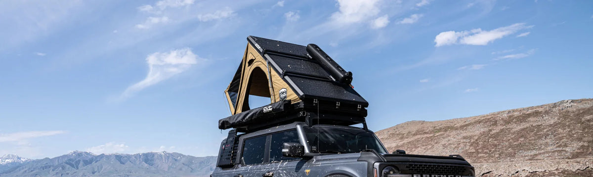 A black SUV with a rooftop tent and solar panels is parked outdoors on rugged terrain, with mountains and a blue sky with clouds in the background.