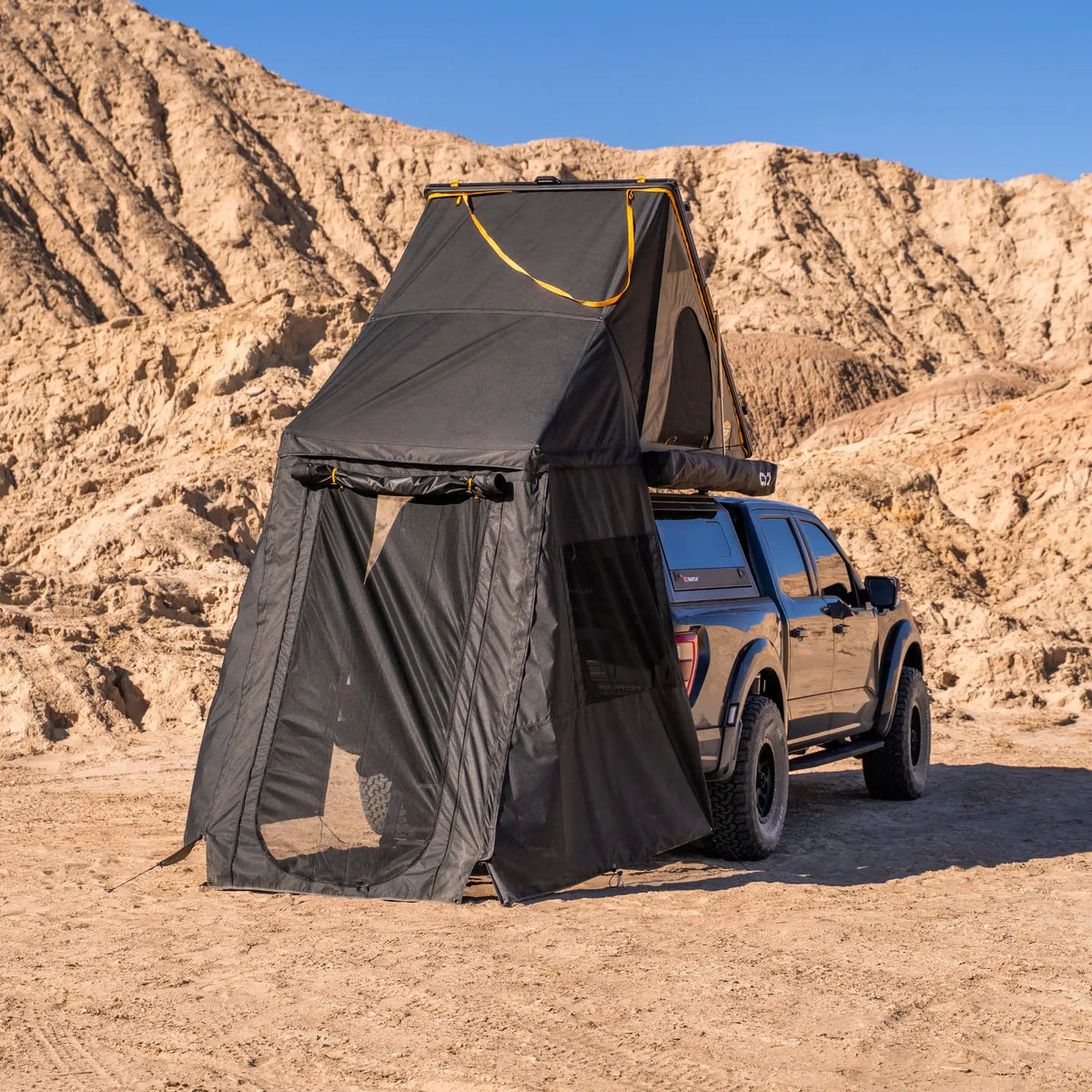 A black pickup truck equipped with a CVT Mt. Hood tent and the CVT Mt. Hood Annex is parked on sandy terrain with rocky hills in the background under a clear blue sky, offering ample outdoor gear storage and enhanced privacy for any adventure.