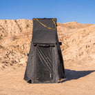 The CVT Mt. Hood Annex, a tall black pop-up privacy tent, stands on sandy ground with tan rocky hills and a clear blue sky in the background—serving perfectly as an outdoor gear storage solution or as a versatile tent annex for added convenience.