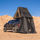 A black pickup truck is parked on sand under a clear blue sky, with a roof tent extended upward and a spacious CVT Mt. Hood Annex attached, providing ample outdoor gear storage.