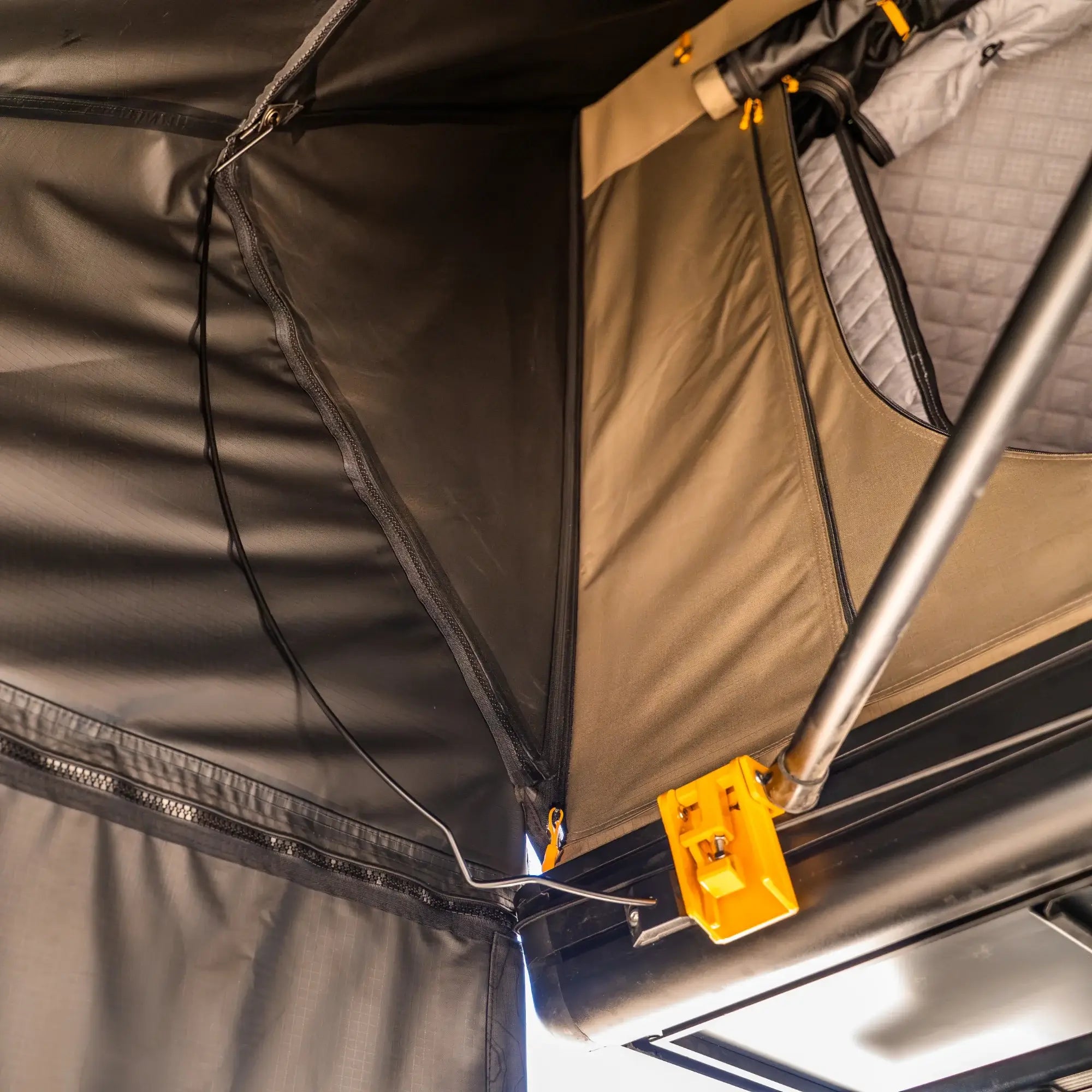 Close-up of an open rooftop tent with black and tan fabric, visible support poles, wires, and a yellow latch mechanism attached to the frame. The tents interior lining and zippered opening are partially visible.