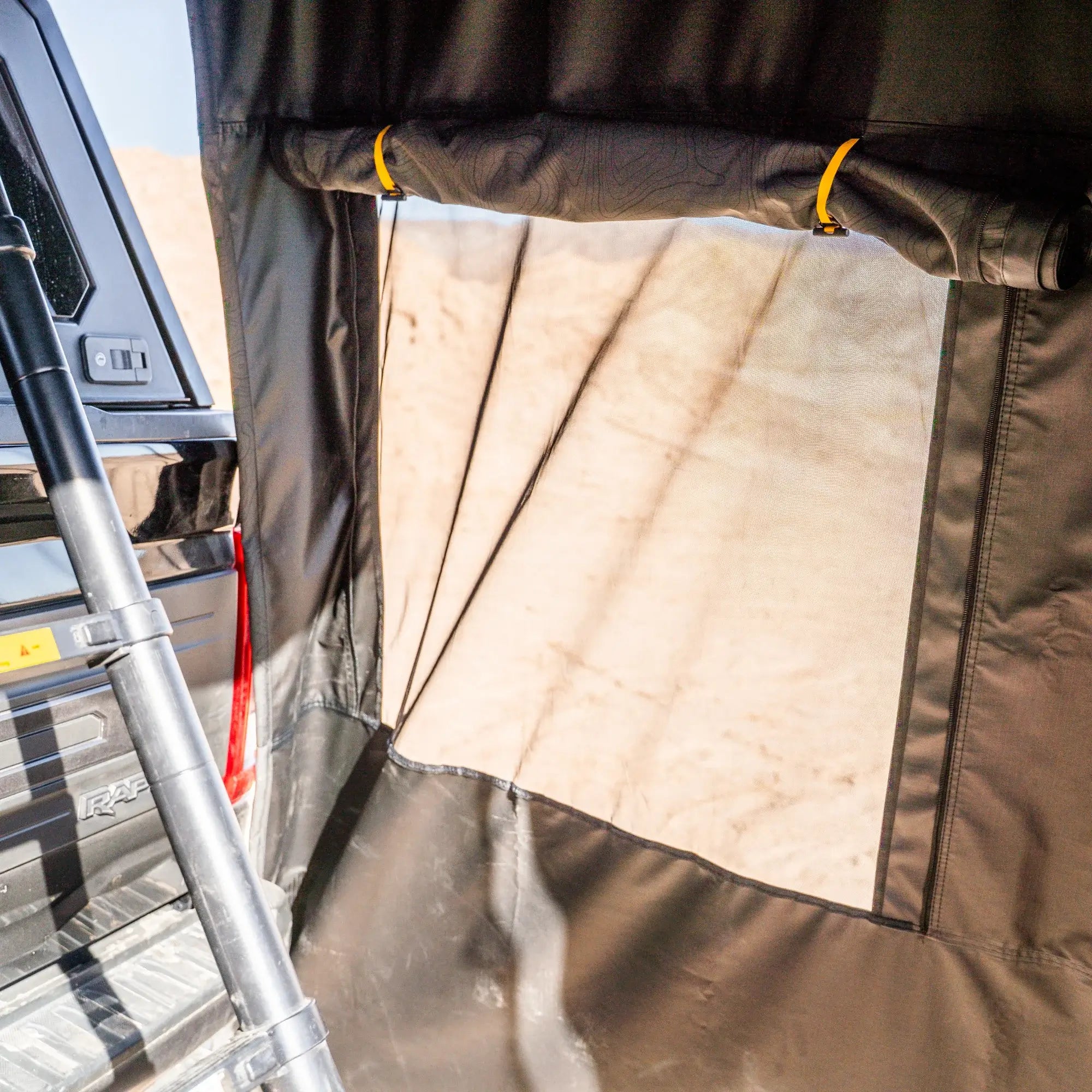 A mesh window with a rolled-up cover and yellow ties is shown inside a tent attached to a truck bed, providing a view of sandy terrain outside.