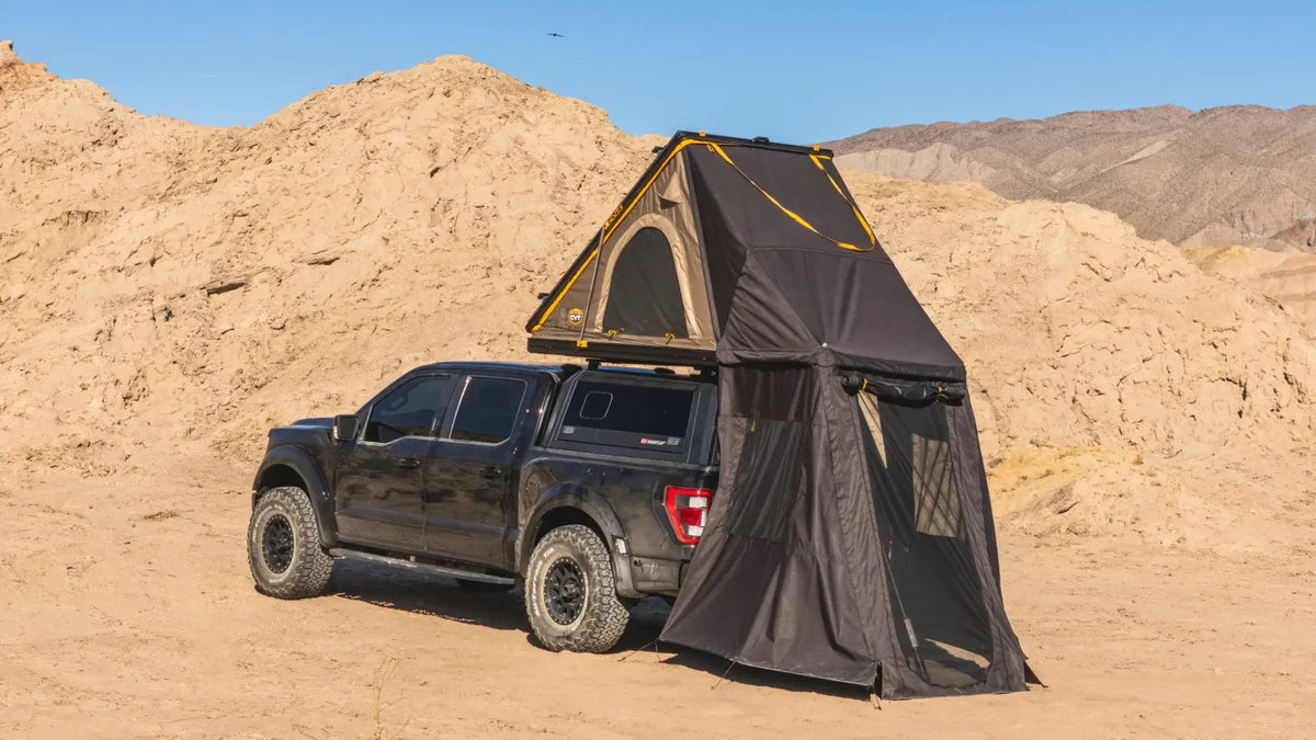 A black pickup truck with a rooftop tent and attached privacy enclosure is parked in a sandy desert area with hills in the background under a clear blue sky.