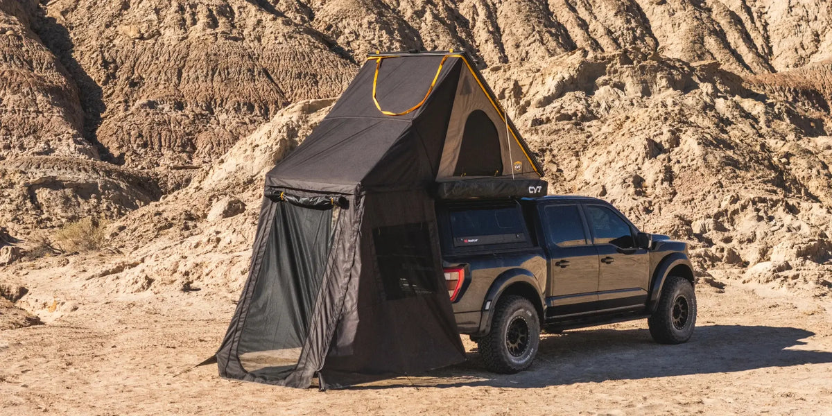 A black pickup truck is parked in a rocky desert, equipped with a rooftop tent and attached ground-level shelter with mesh walls, creating a two-level camping setup.