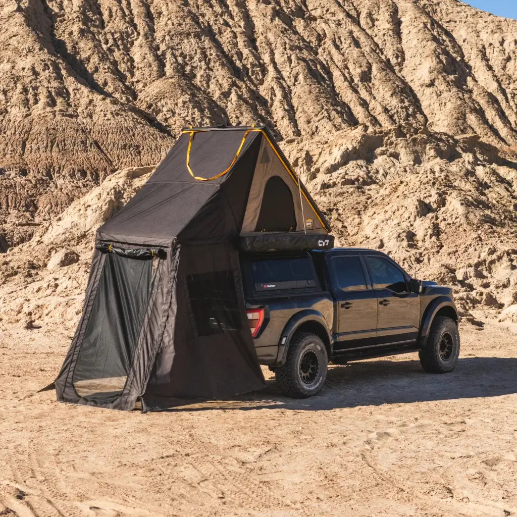 A black pickup truck is parked in a desert landscape with a rooftop tent extended, including a mesh annex area on the ground. Rocky sand dunes form the background.