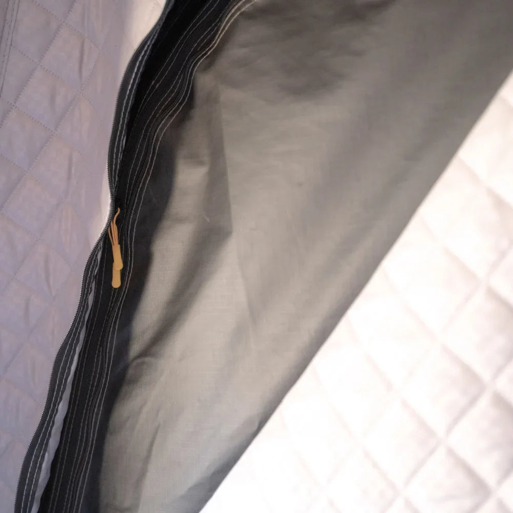 A close-up of a partially unzipped black zipper on gray fabric, bordered by quilted white panels, showcases the CVT Mt. Hood Insulation Walls—an insulated liner engineered to boost tent warmth during cold-weather camping.