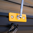 A close-up of a metal latch attached to a yellow plate with two screws, mounted on a dark surface. A metal rod is secured in the latch, and a black carabiner hangs below—an essential feature for securing gear during outdoor adventures or when setting up your CVT Mt. Hood 2 Hardshell Tent.