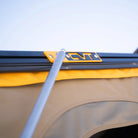 A close-up of a tent pole connected to a black metal frame with a yellow CVT bracket, with tan and yellow fabric from the Mt. Hood 2 Hardshell Tent by CVT visible underneath and a clear sky in the background—showcasing the durability and thoughtful design that make this rooftop tent ideal for your outdoor adventures.