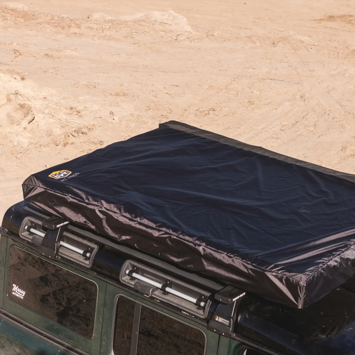 A CVT Mt. Hood 2 Hardshell Tent in black, securely enclosed in its cover, is mounted atop a green vehicle parked on sandy desert terrain—an ideal setup for outdoor adventures.