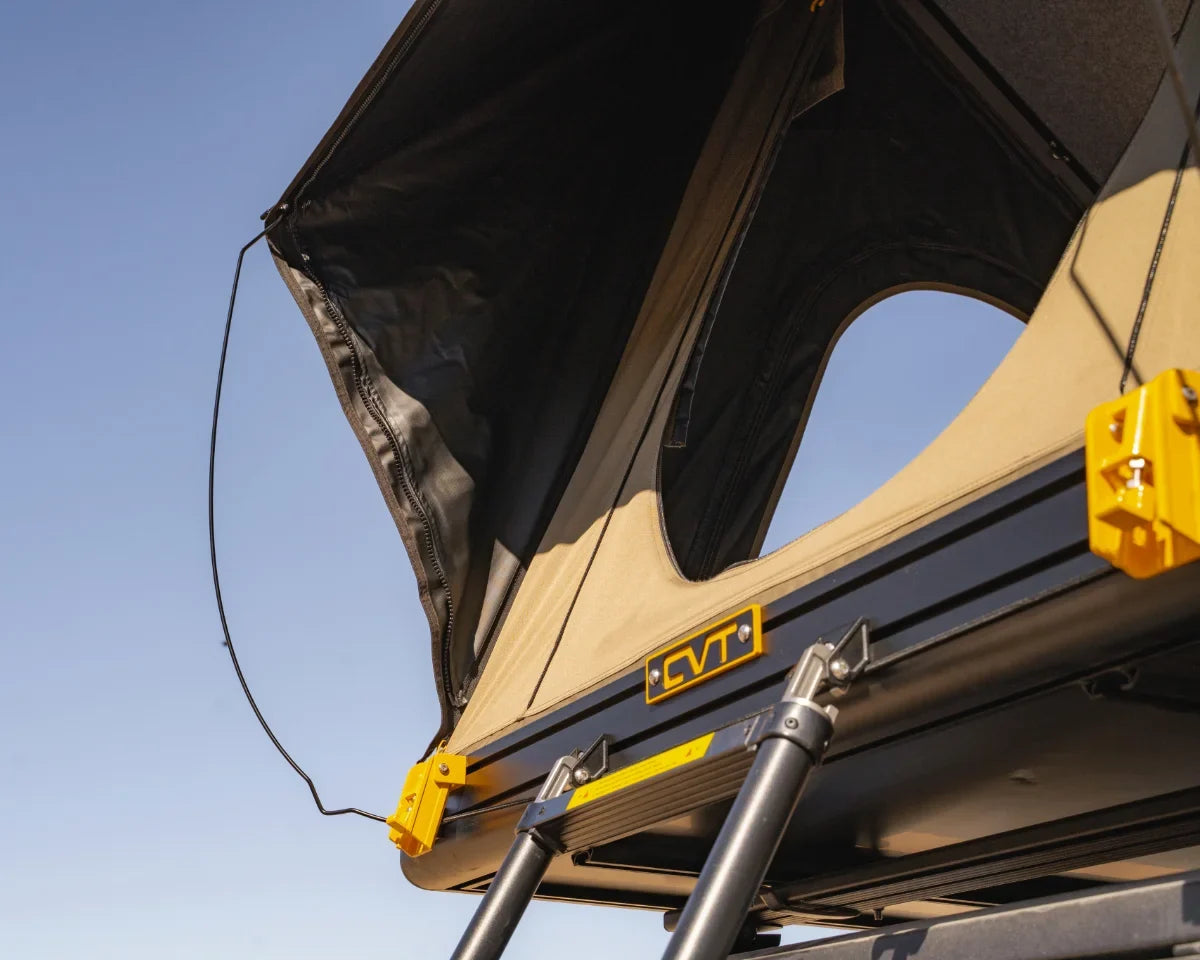 A rooftop tent with an open canvas cover and ladder is mounted on a vehicle against a clear blue sky. The tent features a CVT logo and yellow corner brackets.
