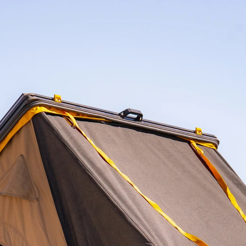 A close-up of the corner of a rooftop tent with yellow straps and a black hard shell, featuring the CVT Mt. Hood Handle for a secure grip, set against a clear blue sky.