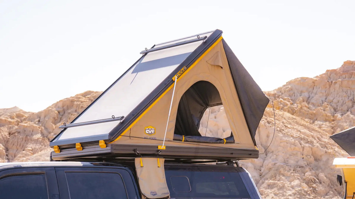 A rooftop tent is set up on top of a vehicle parked in a rocky, desert-like area. The tent is tan with black and yellow accents, featuring an open side window and a small hanging pouch.