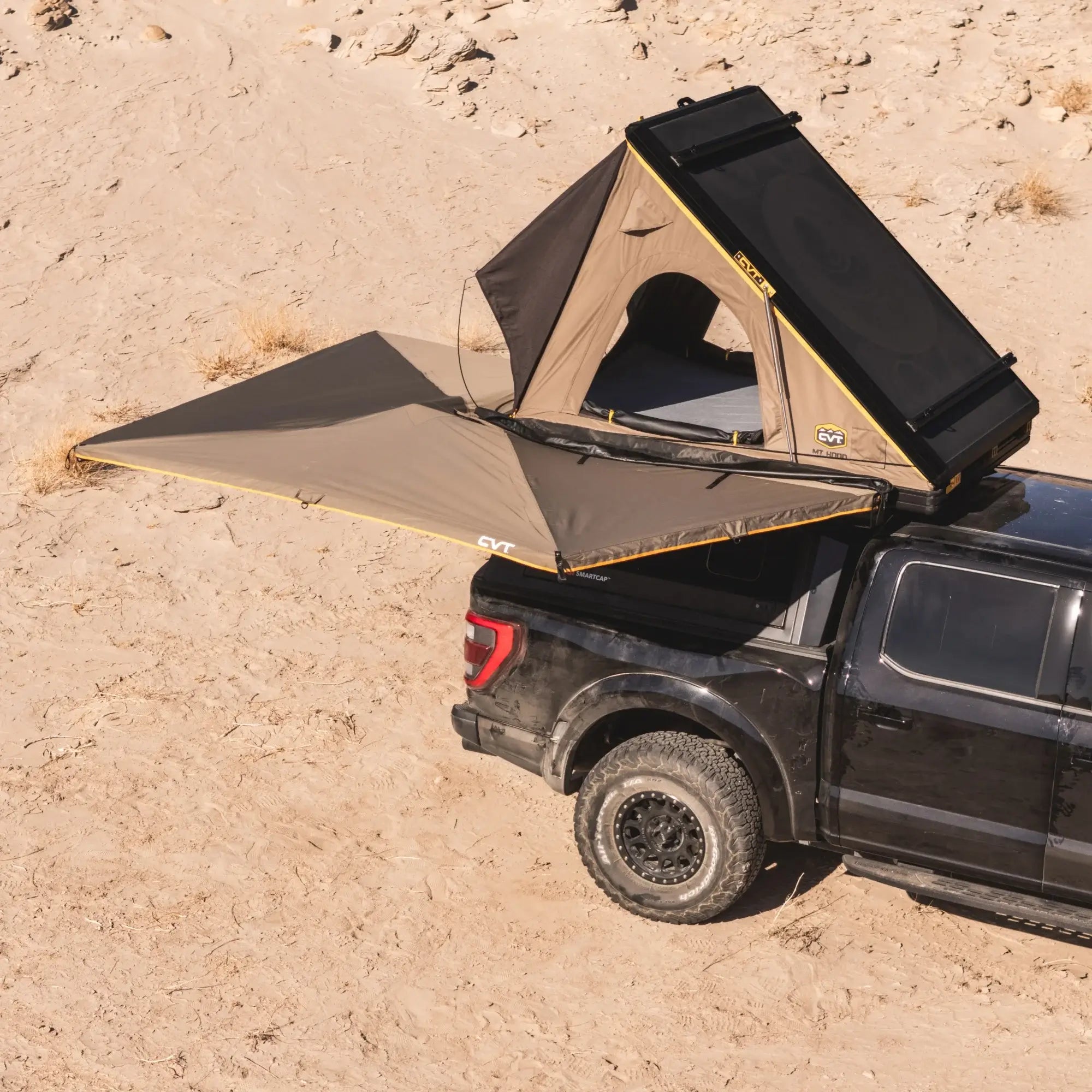 A black pickup truck with a rooftop tent and an extended awning is parked on sandy terrain, ready for camping in a desert environment.