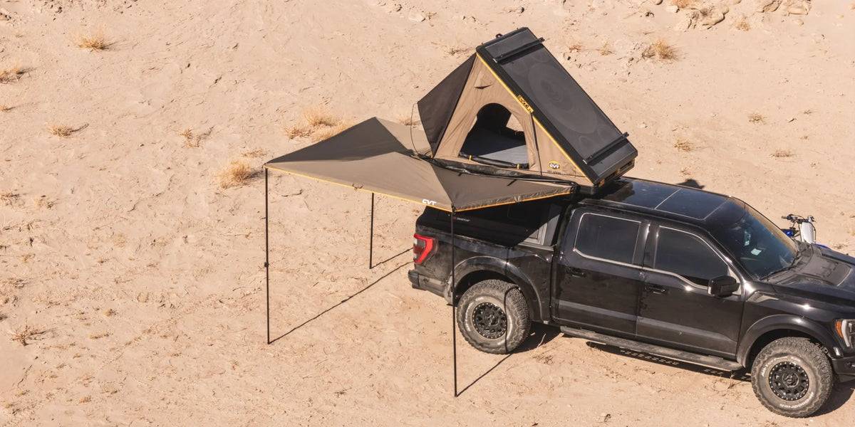 A black pickup truck in the desert with a rooftop tent and an extended awning attached, providing shade and shelter. The sand around is light and dry with minimal vegetation.