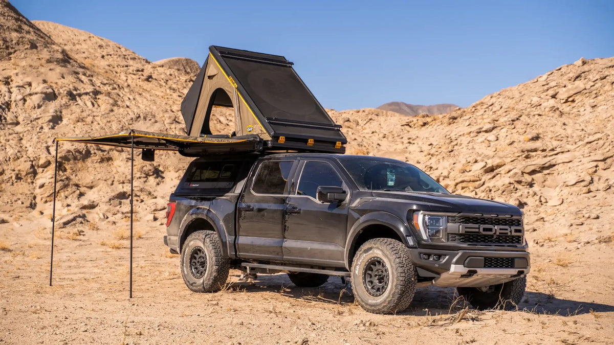 A black pickup truck with off-road tires is parked in a rocky desert. It has a rooftop tent set up and an awning extended for shade, with hills and blue sky in the background.