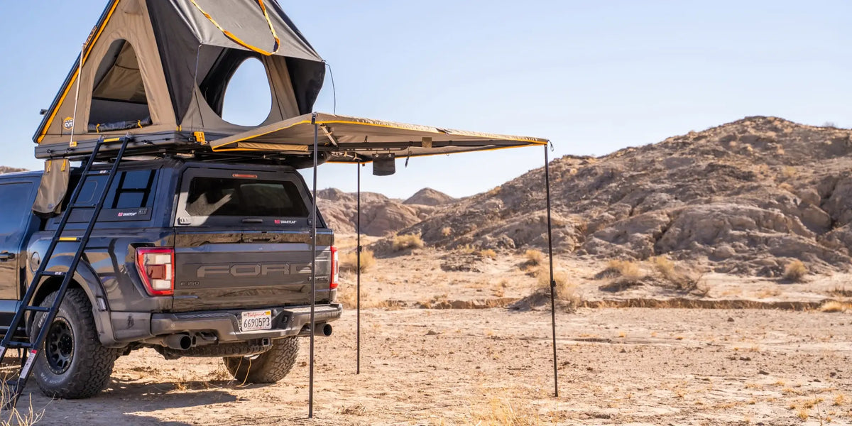 A black pickup truck with a rooftop tent and awning is parked in a dry desert landscape with rocky hills in the background. A ladder leads up to the tent, and the awning provides shade at the rear of the truck.