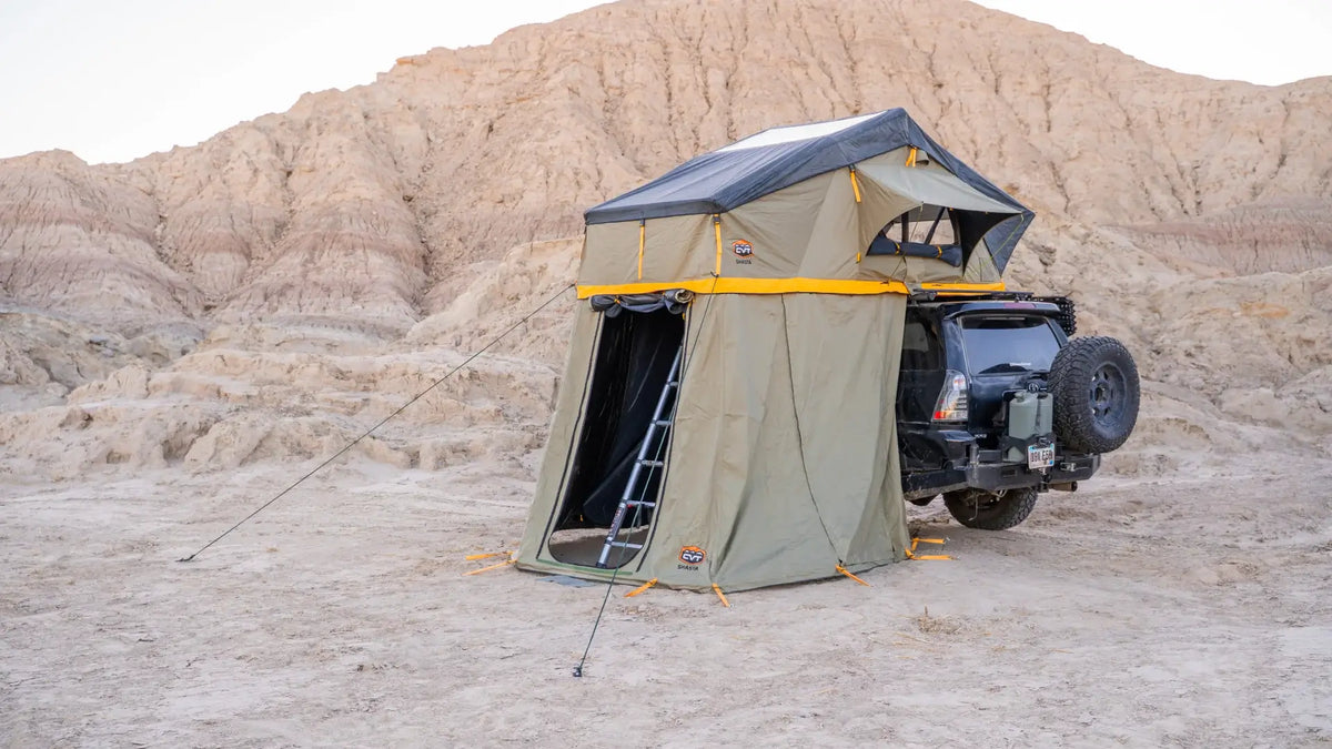 A rooftop tent with an annex is set up on a black SUV, parked on sandy terrain with rocky hills in the background. A ladder connects the tent to the ground.