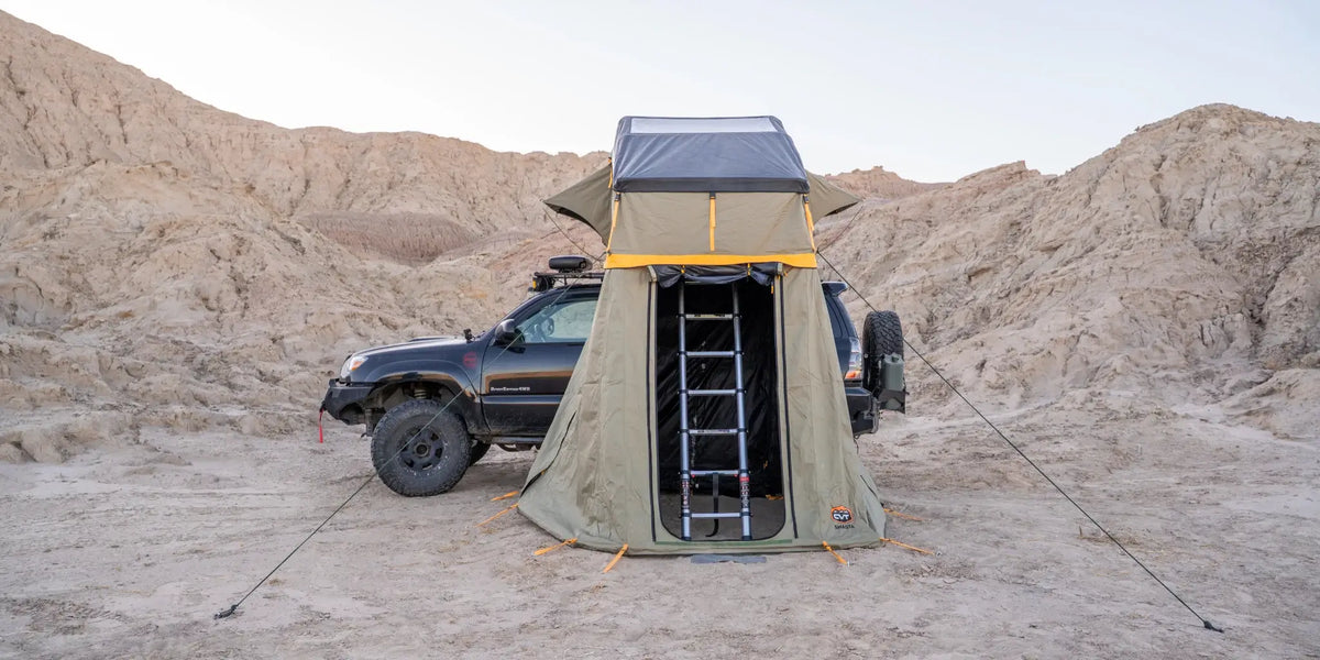 A black off-road vehicle with a rooftop tent and attached ladder is parked in a sandy desert area with rocky hills in the background under a clear sky.