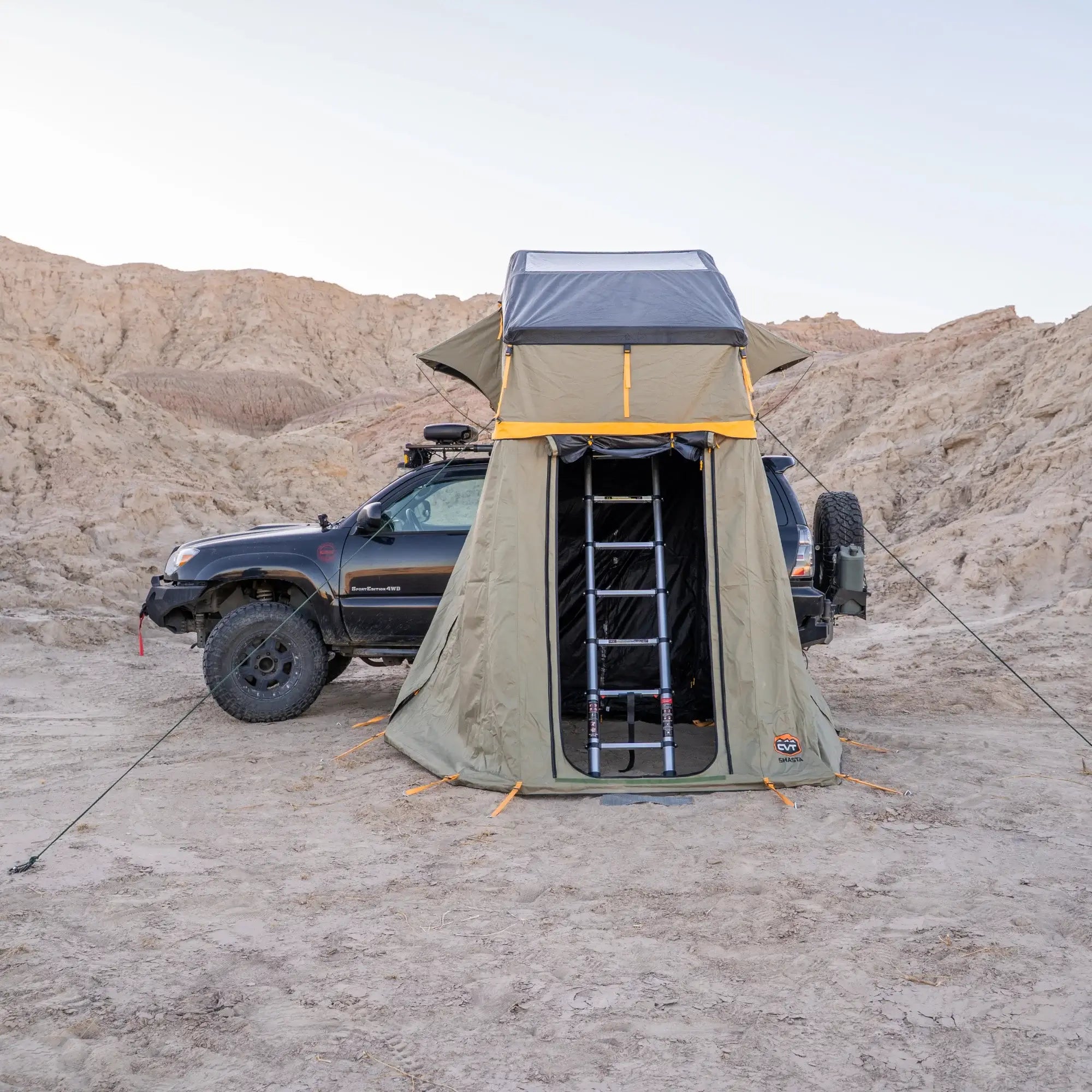 A black SUV with a rooftop tent and ladder is parked on sandy desert terrain surrounded by rocky hills under a clear sky, featuring added comfort from the CVT Pioneer Annex, which provides an enclosed tent space.