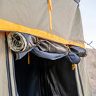 Close-up of a rolled-up tent flap secured with ties, showcasing the yellow and gray fabric and visible zipper of the CVT Pioneer Annex, with part of its extension entrance and sky in the background, offering a glimpse into the enclosed tent space.