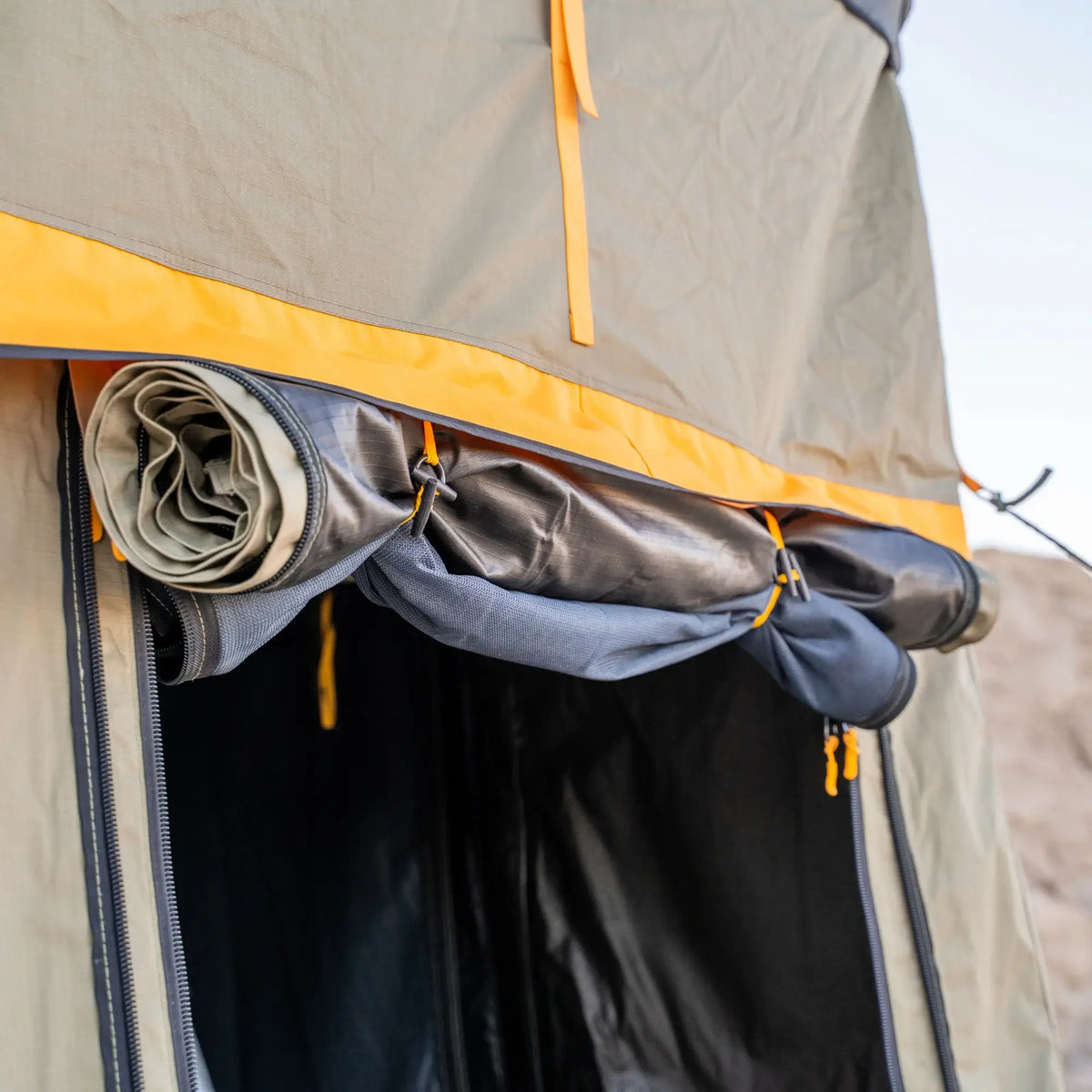 Close-up of a rolled-up tent flap secured with ties, showcasing the yellow and gray fabric and visible zipper of the CVT Pioneer Annex, with part of its extension entrance and sky in the background, offering a glimpse into the enclosed tent space.