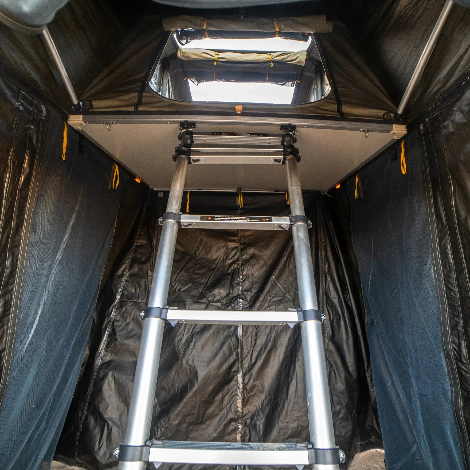 View from inside an enclosed tent space, looking up at a metal ladder leading to the rooftop tent opening, with dark fabric walls on both sides—the ideal setup for attaching your CVT Pioneer Annex.