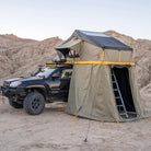 A black off-road vehicle with a rooftop tent and attached awning from CVT is parked on sandy terrain with rocky hills in the background. Equipped with the CVT Pioneer Annex for added enclosed tent space, a ladder leads from the ground up to the elevated tent.