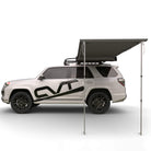 A white SUV with a roof rack and black graphics is parked with a large, rectangular CVT Pioneer Awning - 79″ Medium extended from the roof, supported by two poles, providing versatile outdoor shelter.