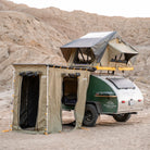 A teardrop camper equipped with CVT Pioneer Awning Walls made from waterproof ripstop material, along with a rooftop tent, is parked in a desert landscape with rocky hills in the background.