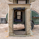 A tan Pioneer Awning Walls tent by CVT with an open front stands in front of a green teardrop camper on sandy terrain, revealing a bed inside. Crafted from waterproof ripstop material, the tent is secured at the corners with orange ties and features mosquito netting for added protection. Camping gear is visible around the camper.
