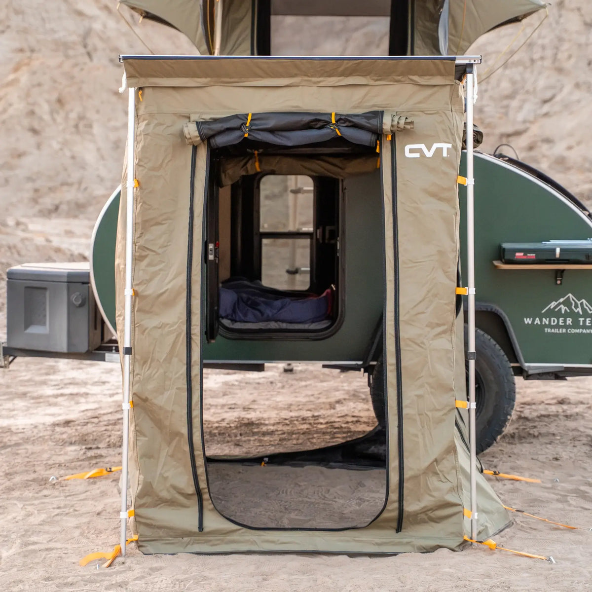A tan Pioneer Awning Walls tent by CVT with an open front stands in front of a green teardrop camper on sandy terrain, revealing a bed inside. Crafted from waterproof ripstop material, the tent is secured at the corners with orange ties and features mosquito netting for added protection. Camping gear is visible around the camper.