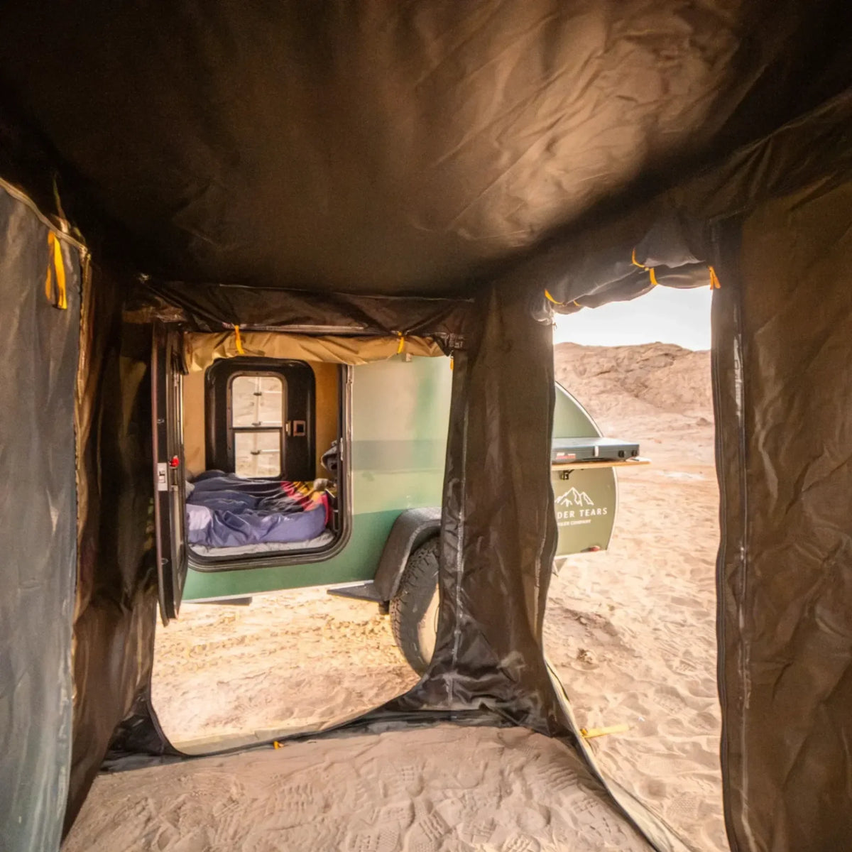 View from inside a CVT Pioneer Awning Walls setup, looking out at a green camper trailer parked on sandy ground, with a bed visible through the trailer door and rocky hills in the background.