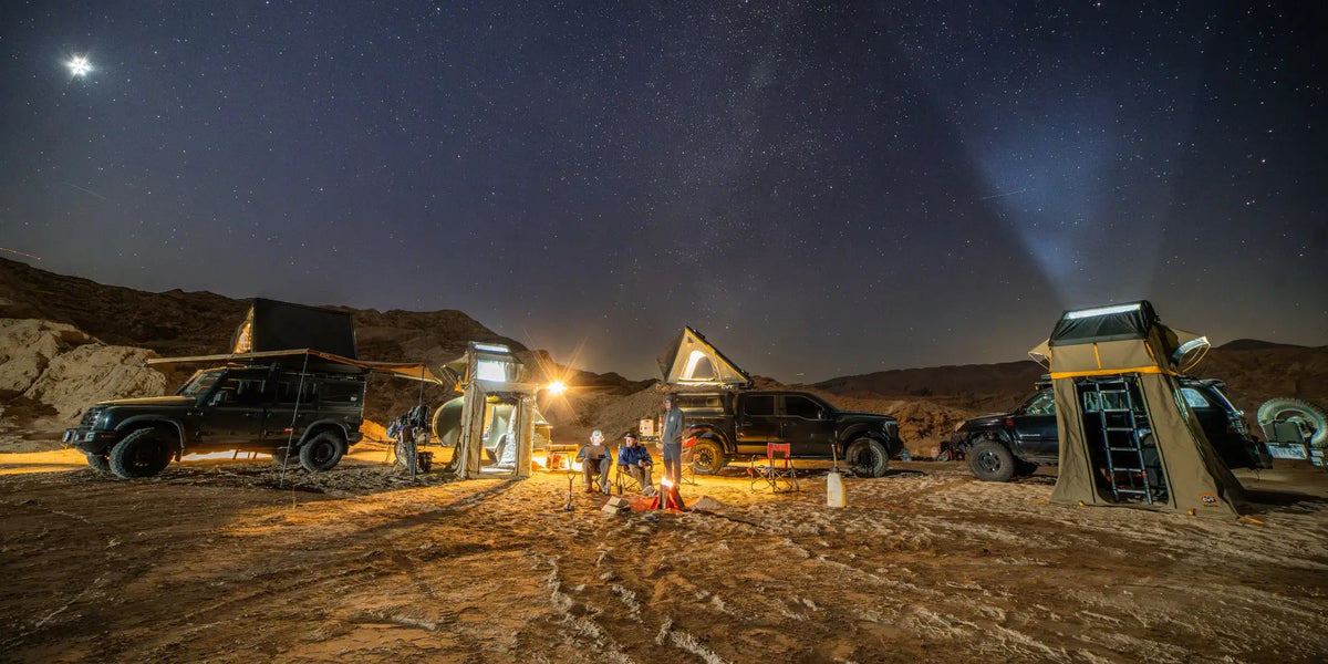 Four off-road vehicles with rooftop tents are parked in a desert under a starry night sky, illuminated by camp lights, with people sitting around a small campfire in the center.