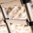 Close-up of the CVT Pioneer Ladder, a metal telescoping ladder featuring black plastic joints, set against a blurred, sandy background.