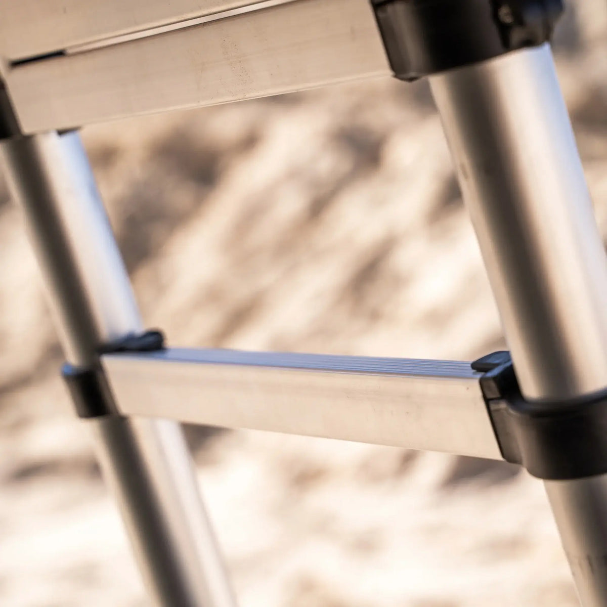 Close-up of the CVT Pioneer Ladder, a metal telescoping ladder featuring black plastic joints, set against a blurred, sandy background.