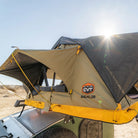 A CVT Pioneer 2 Softshell Tent is set up on a vehicle in a sunny, sandy outdoor setting, with sunlight shining from behind the tent—an ideal setup for overland adventures.