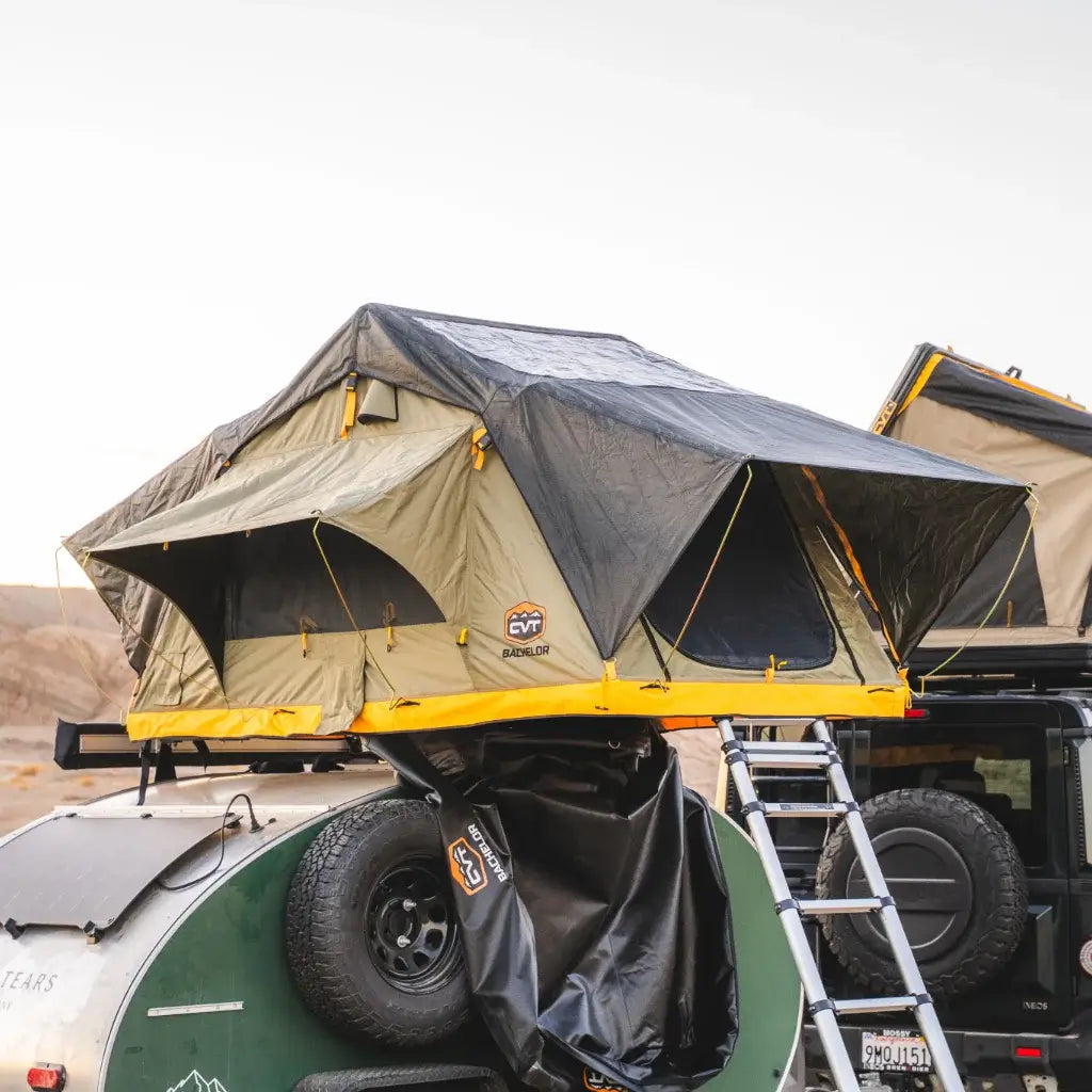 A CVT Pioneer Ladder extends from a rooftop tent with rainfly, set up atop a vehicle beside a compact camper trailer in the desert, offering sturdy and easy access for outdoor adventurers.