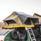 A CVT Pioneer Ladder extends from a rooftop tent with rainfly, set up atop a vehicle beside a compact camper trailer in the desert, offering sturdy and easy access for outdoor adventurers.