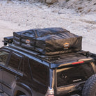 A black SUV with a CVT Pioneer 2 Softshell Tent mounted on a roof rack is parked on rocky, sandy terrain—perfect for overland adventures. The soft-shell tent is folded and secured in a sleek black protective case.