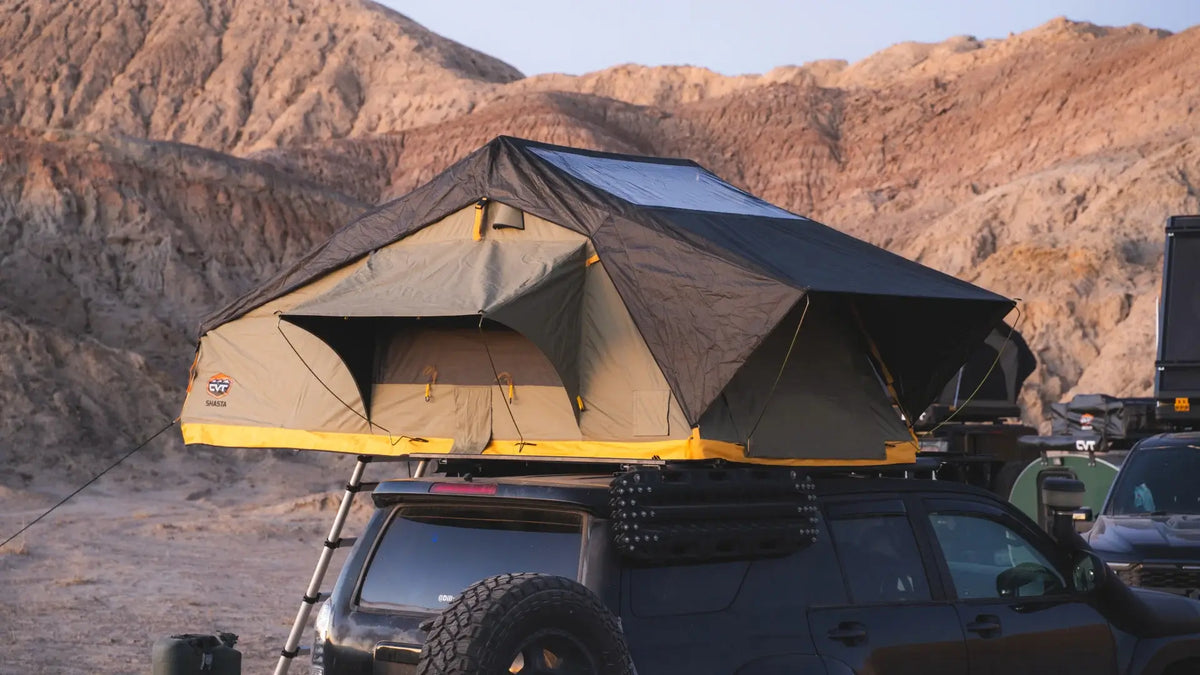 A rooftop tent is set up on top of a black SUV parked in a rocky desert landscape, with tan mountains in the background under a clear sky.