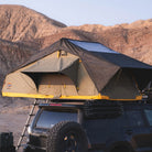 The CVT Pioneer 2 Softshell Tent is mounted on top of an off-road vehicle parked in a rocky desert landscape, with a ladder leading up to the tent—an ideal setup for overland adventures.