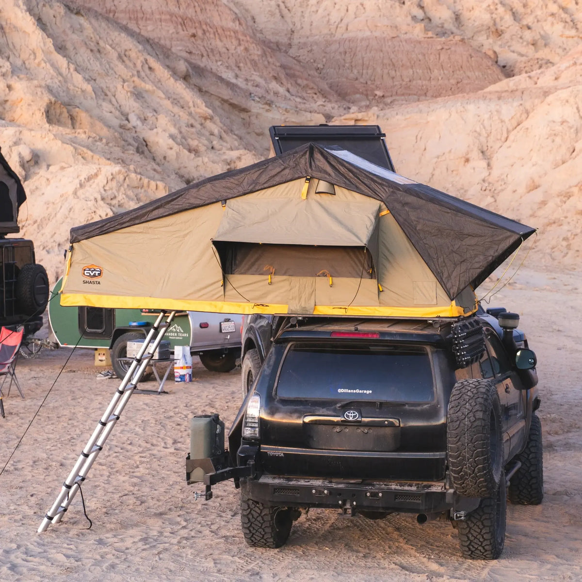A CVT Pioneer 2 Softshell Tent is set up on top of a black SUV parked in a sandy desert area, with a ladder leading up to the tent and rocky hills in the background—perfect for overland adventures.